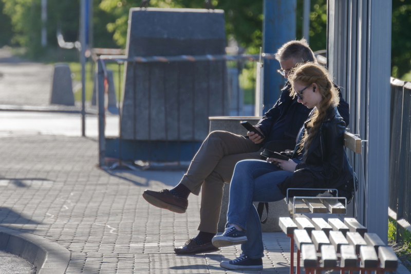 23 - Two people sitting on a bench at a bus station, looking at their mobile devices